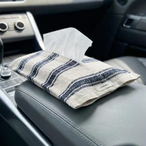 May include: A beige fabric tissue box cover with navy blue stripes, placed on a car's center console. White tissues are partially visible. The cover has a rectangular shape and a small tag with the text "LOVIN&ANIA".