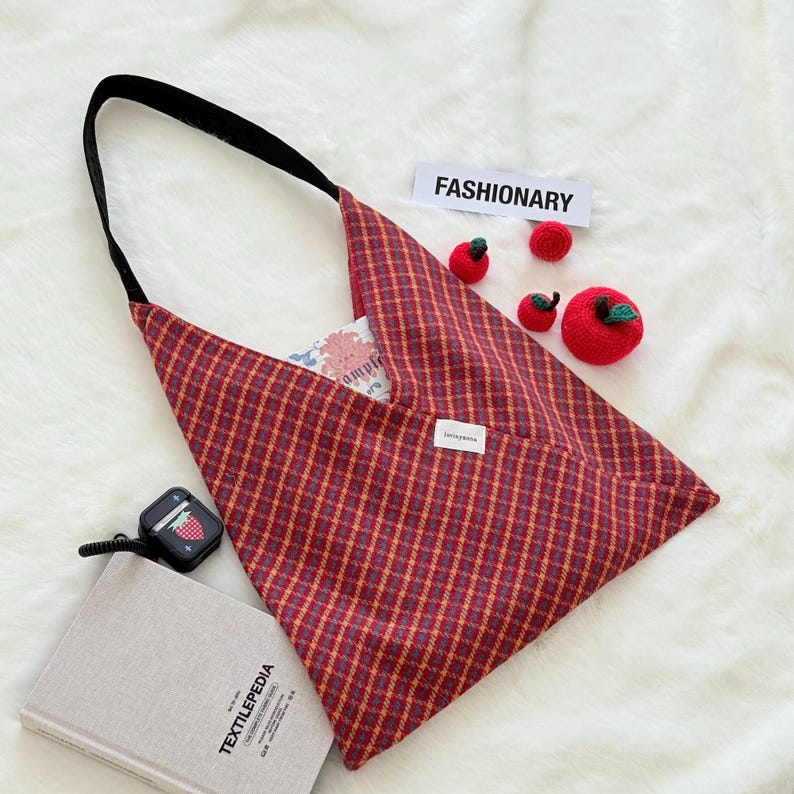 Puede incluir: Un bolso tote a cuadros rojos y dorados con una correa negra. El bolso tiene forma triangular y una peque&ntilde;a etiqueta blanca. La palabra "FASHIONARY" est&aacute; en una etiqueta blanca. Tambi&eacute;n hay varias manzanas rojas de ganchillo.