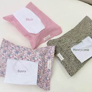 Pode incluir: Três capas retangulares de tecido para lenços. Uma é rosa sólida com a etiqueta Blush, uma apresenta um padrão de favo de mel verde e branco com a etiqueta Honeycomb, e uma exibe uma estampa floral de coelhos rosa e azul com a etiqueta Bunny.