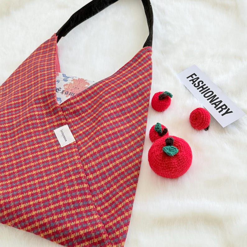 Puede incluir: Un bolso tote a cuadros rojos y amarillos con una correa negra y varios adornos de manzana roja de ganchillo con hojas verdes. La palabra "FASHIONARY" est&aacute; impresa en una tarjeta blanca.