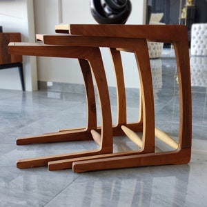 Könnte beinhalten: Drei verschachtelte Holztische mit einem geschwungenen Design. Die Tische sind aus hellem braunem Holz gefertigt und übereinander gestapelt.
