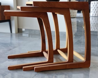 May include: Three wooden nesting tables with a curved design. The tables are made of a light brown wood and are stacked on top of each other.