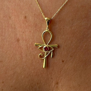 Pode incluir: Um colar com pingente dourado apresentando um símbolo Ankh com um design de Olho de Hórus. O pingente tem uma pequena pedra preciosa vermelha e redonda no centro do olho. O colar é usado ao redor do pescoço.