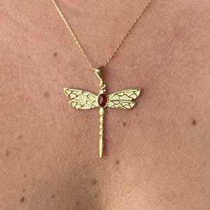 以下が含まれることがあります： 金色のトンボペンダントネックレス。 トンボは質感のある羽と赤い宝石の中心を持っています。 ネックレスは繊細なゴールドチェーンに付いています。