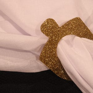 May include: A gold glitter napkin ring in the shape of a house. The ring is on a white linen napkin.