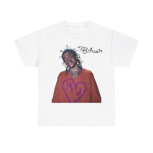 Puede incluir: Camiseta blanca con un gráfico de una persona con un diseño en forma de corazón en su suéter. El texto "Tezo Touchdown" está escrito sobre el gráfico.