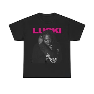 May include: Black t-shirt featuring a grayscale image of a person with dreadlocks, wearing a black hoodie. The word "LUCKI" is printed in bright pink across the top. The hoodie has ornate lettering on the sleeve.