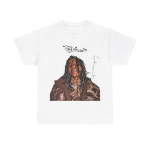Puede incluir: Camiseta blanca con un gráfico de una persona con el pelo largo que lleva una chaqueta marrón y de camuflaje. El texto "Tezo Touchdown" está impreso en negro sobre el gráfico.