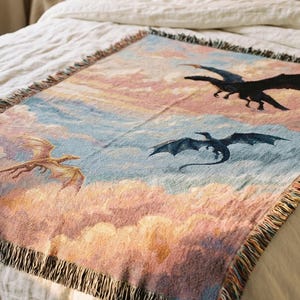 Könnte beinhalten: Eine gewebte Wurkdecke mit einer Fantasieszene mit Drachen, die durch einen pastellfarbenen Himmel fliegen. Die Decke hat einen Fransenrand und ist über einem Bett mit weißer Bettwäsche drapiert. Die Drachen sind in Schwarz-, Blau- und Goldtönen gehalten.