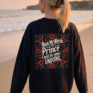May include: Black sweatshirt with the text "Mark My Words Prince I will be your UNDOING" surrounded by red flowers and green leaves. The person is standing on a beach.