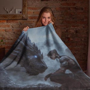 May include: A soft, blue fleece blanket featuring a graphic print of a dragon and a person. The dragon is black and blue with glowing eyes. The person is wearing a dark shirt and trousers and is looking at the dragon.