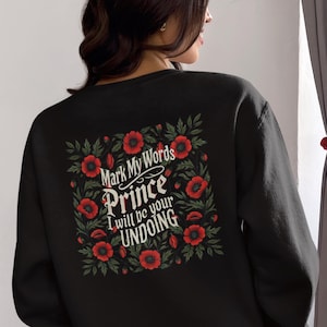 Op de afbeelding: Zwart sweatshirt met een bloemenontwerp en de tekst "Mark My Words Prince I Will Be Your Undoing" gedrukt op de achterkant.