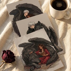 May include: Three art prints featuring fantasy illustrations. One shows a couple embraced with large black wings, another a man with a wolf, and the third a woman reading with a wolf. A cup of coffee and a dried rose are also visible.