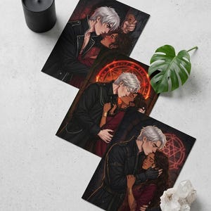 May include: Three art prints depicting romantic illustrations of a couple embracing. The man has white hair and wears a black jacket. The woman has dark hair and wears a red dress. A black candle and a monstera leaf are also present.