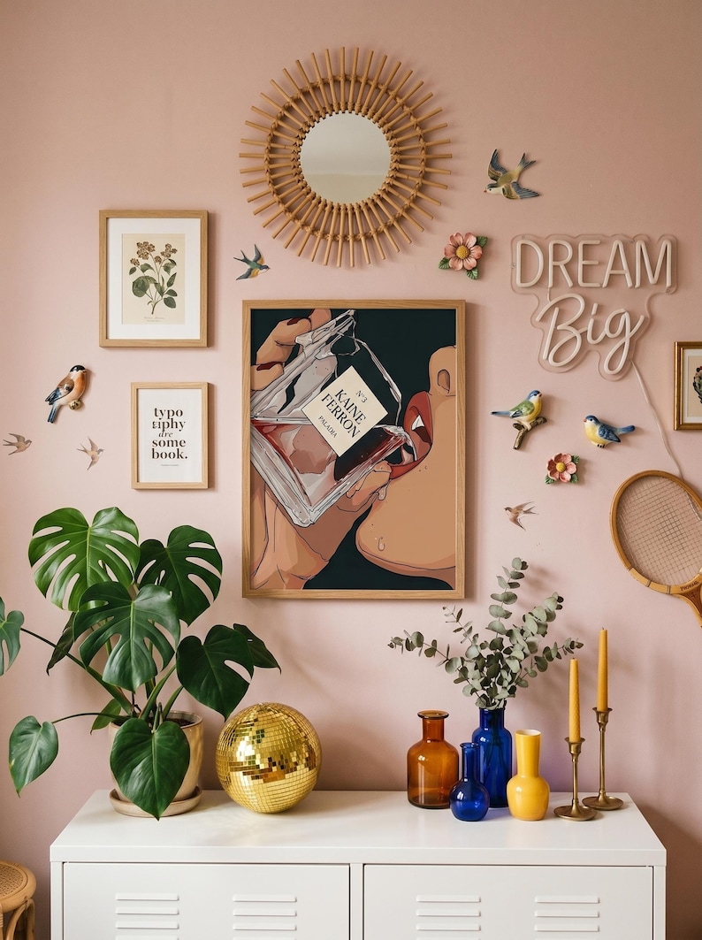 May include: Aesthetic wall decor featuring a framed print of a person drinking perfume, a round mirror, and a neon "Dream Big" sign. Decorative elements include faux birds, flowers, and a disco ball. The color scheme is primarily pink, gold, and green.