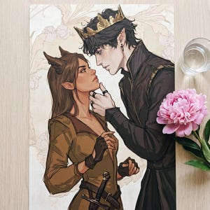 May include: An art print featuring a couple in a fantasy setting. The male figure wears a gold crown and dark clothing, while the female figure has horns and a brown outfit. A pink peony and a glass of water are in the background.