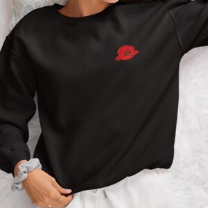 May include: Black sweatshirt with a red embroidered poppy flower on the left chest.
