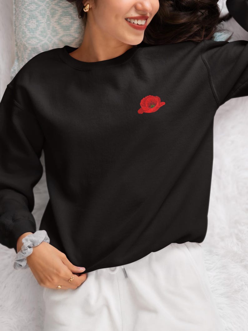 May include: Black sweatshirt with a red embroidered poppy flower on the left chest.