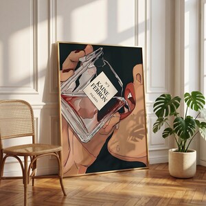May include: A framed art print featuring a stylized illustration of a perfume bottle labeled "KAINE FERRON PALADIA" being held near a person's lips. The artwork has a dark background and a gold frame, with a neutral-toned interior setting.