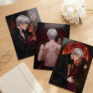 May include: Three art prints with romantic illustrations of a couple. The prints show a man with white hair and a woman with dark hair in different embraces. The prints are on a wooden surface with a white flower and a notebook.