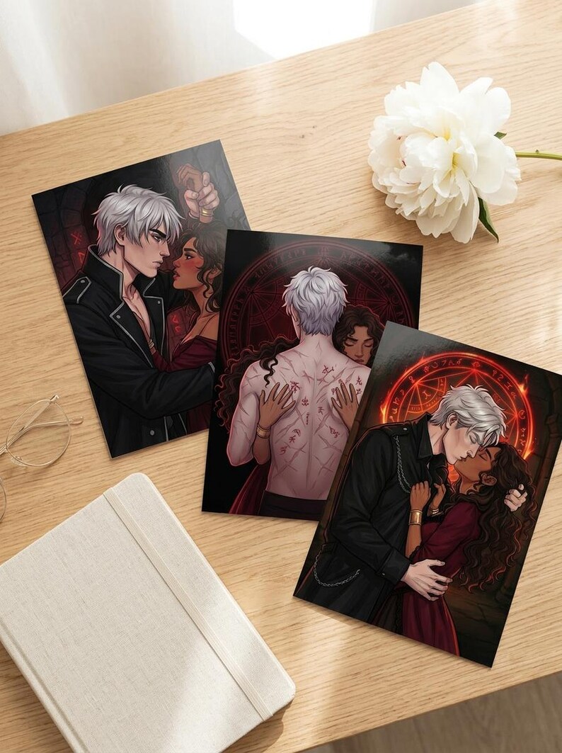 May include: Three art prints with romantic illustrations of a couple. The prints show a man with white hair and a woman with dark hair in different embraces. The prints are on a wooden surface with a white flower and a notebook.