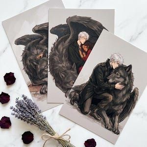 May include: Three art prints featuring illustrations of a wolf with feathered wings, and a person with white hair. One print shows a person embracing the wolf. The prints are on a light gray background. Dried lavender and rose petals are scattered around the prints.