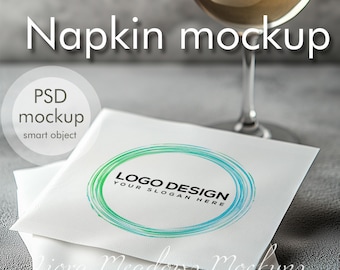 May include: A white napkin mockup with a green and blue circle logo that says "LOGO DESIGN YOUR SLOGAN HERE". The napkin is on a gray surface with a glass of white wine in the background.