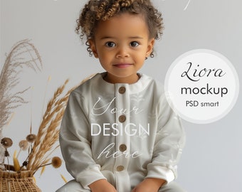 May include: A white onesie with a floral pattern and the text "Your Design Here" on the front. A young child is wearing the onesie and sitting on a wicker stool. The background is a light gray.
