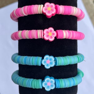 May include: Four bracelets featuring colourful flower charms. The bracelets are made of small, round beads in shades of pink, white, and blue. The flower charms are pink, white, and blue.