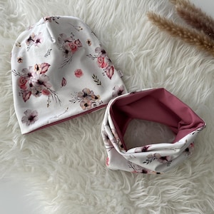 Può includere: Un berretto bianco con stampa floreale e fodera rosa, abbinato a un scaldacollo floreale rosa e bianco.