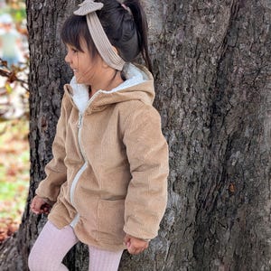 Könnte beinhalten: Ein Kind trägt eine beige Cordjacke mit weißem Fleecefutter und einem passenden Stirnband. Die Jacke hat einen Reißverschluss und Taschen. Das Kind trägt außerdem rosa Strumpfhosen und hellfarbene Stiefel. Warme Kleidung.