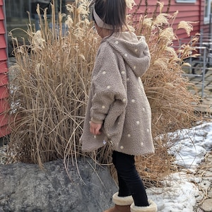 Peut inclure: Une veste à capuche en polaire beige avec un motif de marguerites blanches répétées, portée sur un legging noir et des bottines en daim marron avec une doublure en fausse fourrure crème. La veste présente un tissu texturé type sherpa.