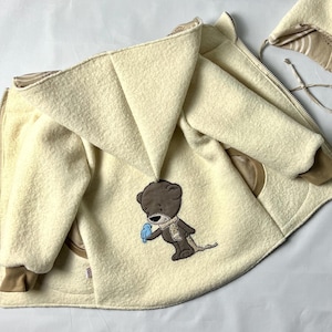 Könnte beinhalten: Eine cremefarbene Kapuzenjacke mit einer Stickerei eines braunen Bären und eines Vogels. Die Jacke hat einen Reißverschluss und eine passende cremefarbene Mütze mit einem braunen Bommel.