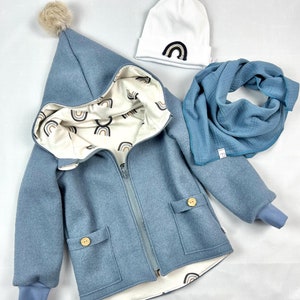 Op de afbeelding: Een blauwe capuchontrui met een witte voering met een regenboogpatroon. De trui heeft een ritssluiting, twee zakken en een pompon op de capuchon. Een witte beanie met een regenboogpatch en een blauwe sjaal zijn ook inbegrepen.