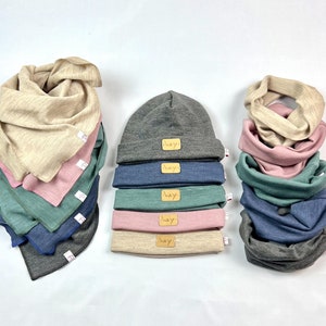 Op de afbeelding: Een set van zes grijze, blauwe, groene, roze en beige nekwarmers met het woord 'hey' erop gedrukt. De nekwarmers zijn op elkaar gestapeld. Er is ook een grijze beanie met het woord 'hey' erop gedrukt.