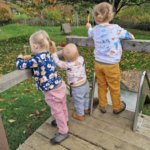 Può includere: Tre bambini giocano su una struttura da gioco in legno. Il bambino in primo piano indossa una camicia a fantasia rosa e blu e pantaloni rosa. Il bambino al centro indossa una camicia a fantasia rosa e bianca e pantaloni blu. Il bambino in fondo indossa una camicia a fantasia bianca e blu e pantaloni gialli.