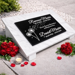 Könnte beinhalten: Schwarze Gedenktafel mit weißem Text und einem Pusteblumen-Design, auf einem weißen Marmorsockel. Die Tafel enthält Namen, Daten und ein Zitat. Rote Rosen und eine Kerze umgeben den Sockel.
