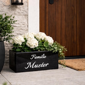 Könnte beinhalten: Eine schwarze, rechteckige Pflanzschale gefüllt mit weißen Hortensienblüten und grünem Laub. Auf dem Pflanzgefäß steht in weißer Schrift "Familie Muster". Die Schale steht auf einer grauen Steinoberfläche.