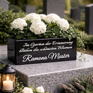 Könnte beinhalten: Ein schwarzer Gedenk-Blumenkasten aus Granit mit weißem Text, der lautet: „Im Garten der Erinnerung blühen die schönsten Blumen Ramona Muster.“ Der Kasten ist mit weißen Hortensien und Grün gefüllt. Eine brennende Kerze in einer Laterne ist im Vordergrund.