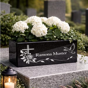 Könnte beinhalten: Ein rechteckiger Pflanzkasten aus schwarzem Granit mit einer eingravierten weißen Kreuzdarstellung, einer Rose und einer fliegenden Taube. Der Name Ramona Muster ist in weißer Schrift eingraviert. Gefüllt mit weißen Hortensien und grünem Laub.