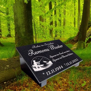 Könnte beinhalten: Ein schwarzer Granitstein mit einer weißen Inschrift, die "Ruhe in Frieden Ramona Muster" und "Spuren im Sand verwehen, Spuren im Herzen bleiben." lautet. Die Inschrift enthält auch die Daten "* 12.11.1954 † 12.11.2022". Der Grabstein zeigt eine Silhouette einer Person beim Angeln.