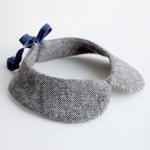 May include: A gray tweed collar with a blue ribbon bow. The collar has a rounded shape and a button closure.
