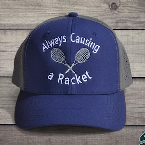 May include: Navy blue baseball cap with a gray mesh back. The cap features the embroidered text "Always Causing a Racket" with crossed tennis rackets. The cap has a curved brim and a button on top.