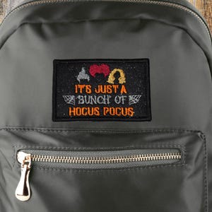 May include: A dark gray backpack with a front zippered pocket and side pockets. The backpack features a patch with the text "IT'S JUST A BUNCH OF HOCUS POCUS" and a logo. The backpack has a top handle and adjustable straps.
