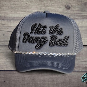 May include: Gray trucker hat with the phrase "Hit the Dang Ball" embroidered in black script. The hat features a mesh back, a curved brim, and a decorative band of rhinestones along the front.