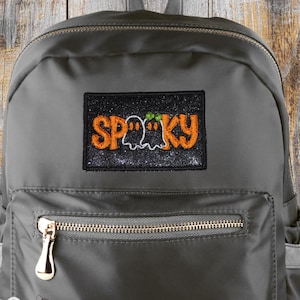 May include: Gray backpack with a front zippered pocket and a top handle. The backpack features a patch with the word "SP00KY" in orange letters, a ghost, and a pumpkin. The backpack has a gold zipper and a tag that says "Stitch with Attitude."