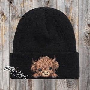 May include: Black beanie featuring a cartoon Highland cow with brown fur, horns, and large eyes. The beanie has a folded cuff and the text "Stitch with Attitude" in white script on the lower left side. The hat is made of a soft, knitted material.
