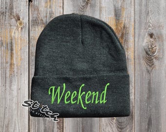 May include: Dark gray beanie with the word "Weekend" embroidered in bright green. The beanie is made of a soft material and is suitable for cold weather. The logo "Stitch with Attitude" is on the lower left.