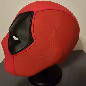 May include: A red Deadpool mask with black eye coverings and a white mesh eye area. The mask is displayed on a black stand, showcasing its textured fabric and distinctive design. A cosplay item.