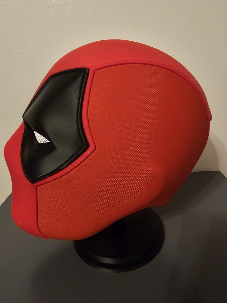 May include: A red Deadpool mask with black eye coverings and a white mesh eye area. The mask is displayed on a black stand, showcasing its textured fabric and distinctive design. A cosplay item.